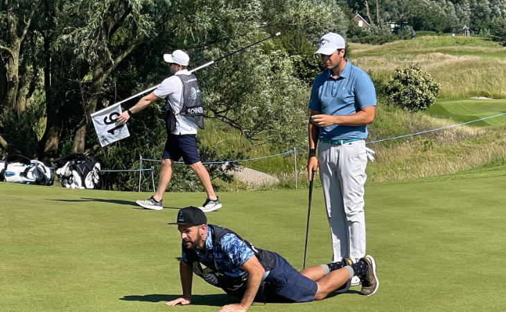 Ein Golfer steht hinter seinem Caddie, der auf dem Rasen liegt und die Puttingline betrachtet