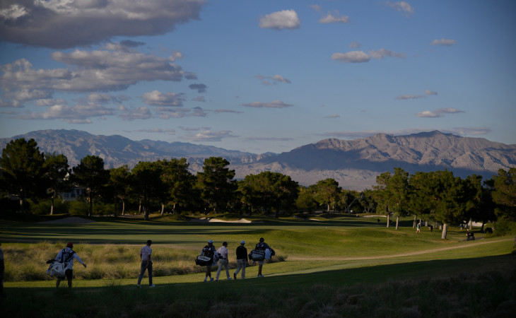 Drei Golfer und ihre Caddies schreiten einen schönen Golfplatz vor einem Bergpanorama entlang.